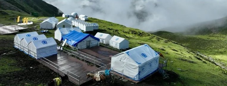 Aerial view of the Siguniang Base Camp: white tents on wooden platforms on a green hillside, with mist rolling in.