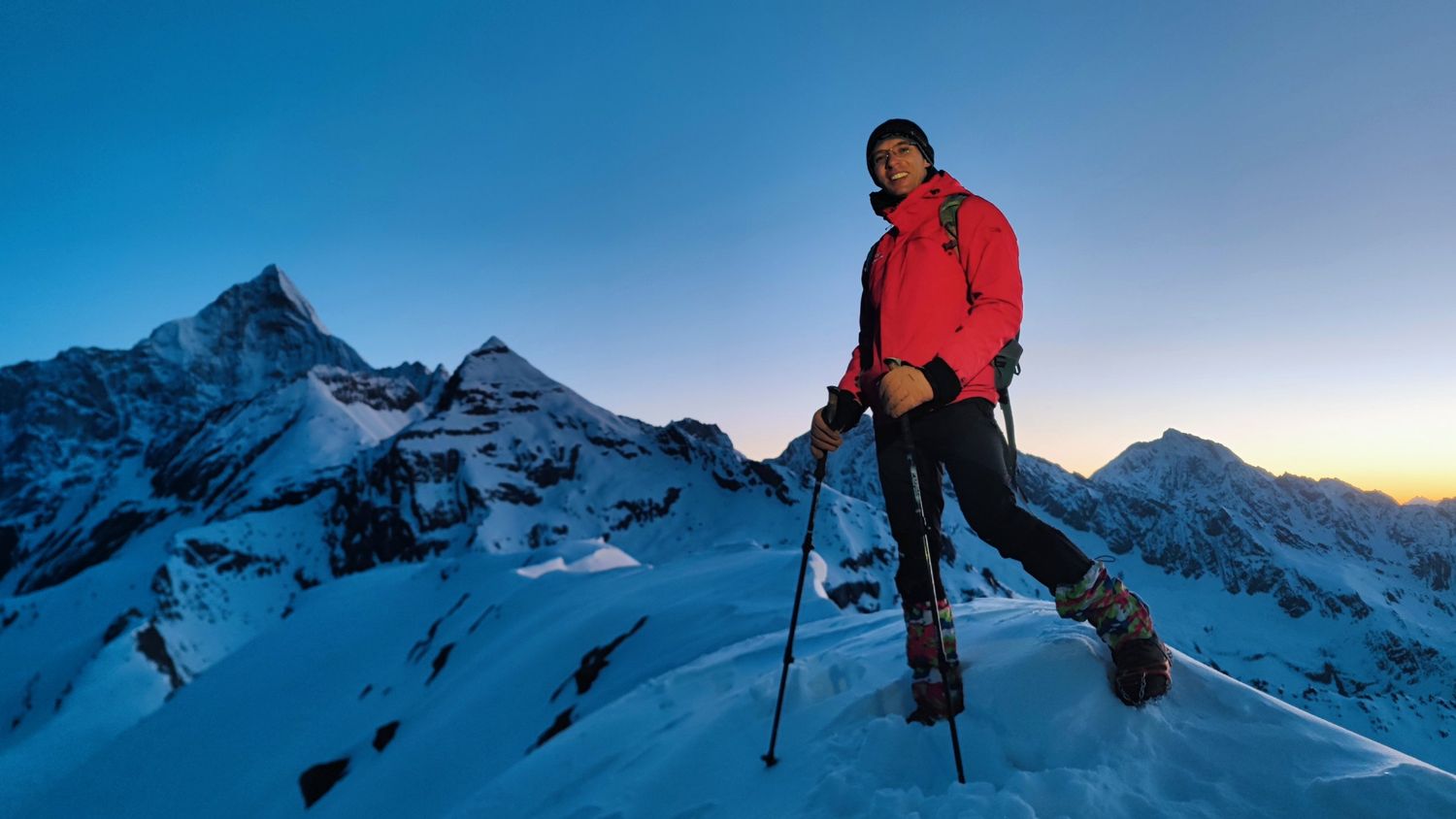 A climber wearing crampons and gaiters smiles on a snowy ridge at dawn, with the peaks of Siguniang behind.