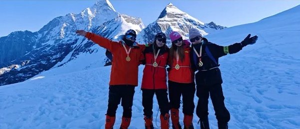 Four climbers in red jackets with arms outstretched on a snowfield, with Siguniang peaks behind them.