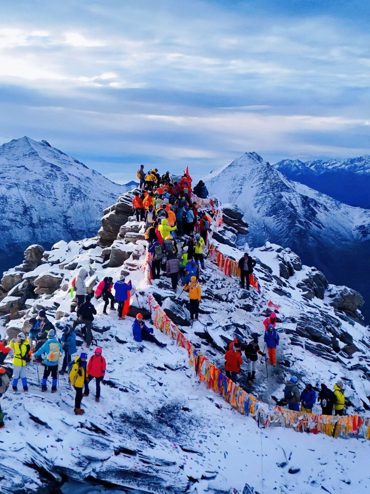 Dozens of climbers in colourful jackets on the snowy summit ridge of Erfeng, with prayer flags, during China's national holiday.