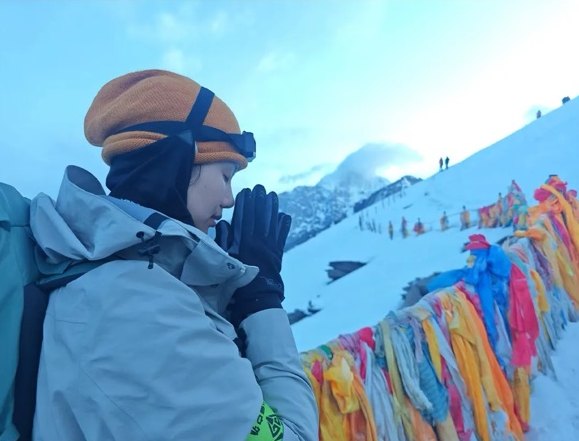 A climber in a beanie and goggles pauses with hands together beside colorful prayer flags on a snowy ridge on Siguniang.