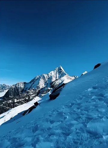 A pointed snow peak of the Siguniang massif seen across a steep blue snow slope under a clear sky.