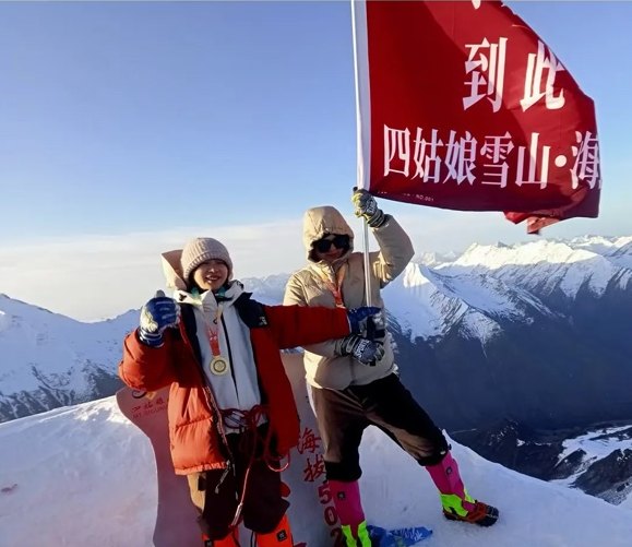 Two climbers hold a red flag at the Siguniang summit with the snowy ranges of Sichuan behind them.