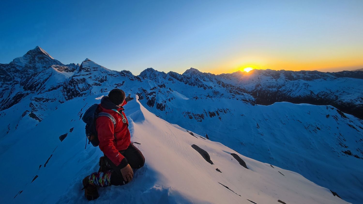 A climber on a snow ridge points toward the sunrise rising over the mountains near Siguniang.